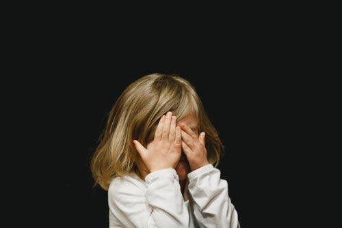 a little girl in fear facing the camera with her eyes covered by her hands over a black background