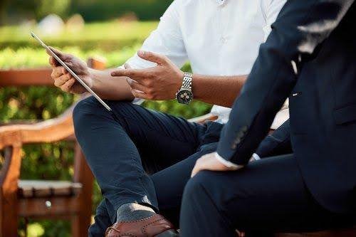 a closeup of a two business men in suits talking on a wooden bench as one man points to his tablet