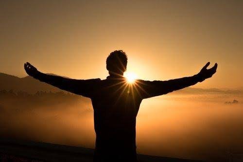 a man's silhouette with outstretched arms in the distance in front of a sunset as the sun's rays peek through