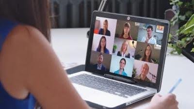 over the shoulder closeup view of a laptop on a table while a female watches participants of the video conference on screen